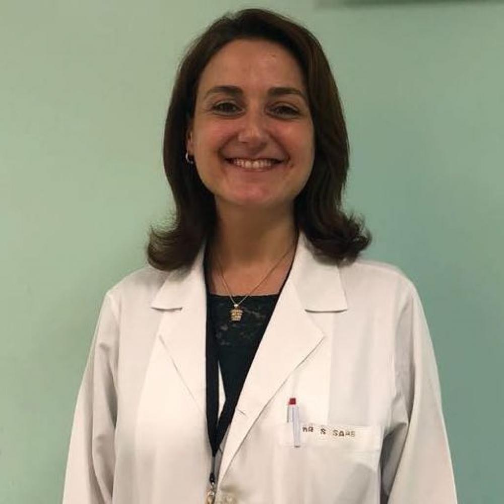 Headshot of Dr. Raya Saab against a mint-green wall background. She is wearing a white coat and has shoulder-length dark-brown hair. She is smiling facing forward.