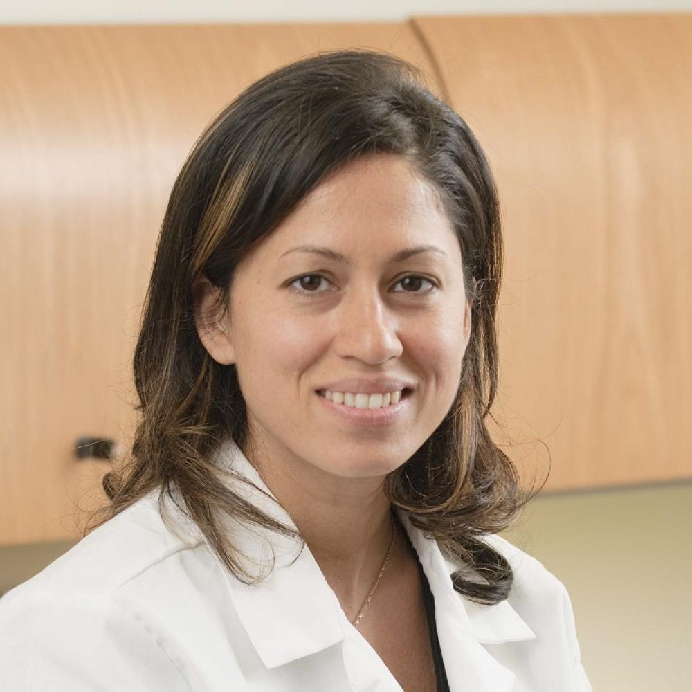 Headshot of Dr. Shanu Modi in a clinical setting wearing a white coat and shoulder-length brown hair.