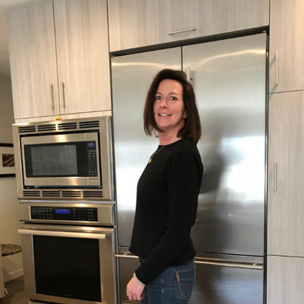 Molly standing in a kitchen. She is smiling facing forward with her body facing sideways. She has shoulder-length brown hair and is wearing a long-sleeve black shirt and jeans.
