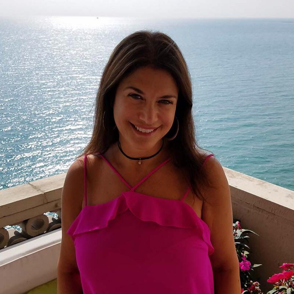 Lillian Kreppel wearing a pink shirt and shoulder-length brown hair against a vast blue-sea background. She is smiling facing forward on a balcony with the sea behind her.