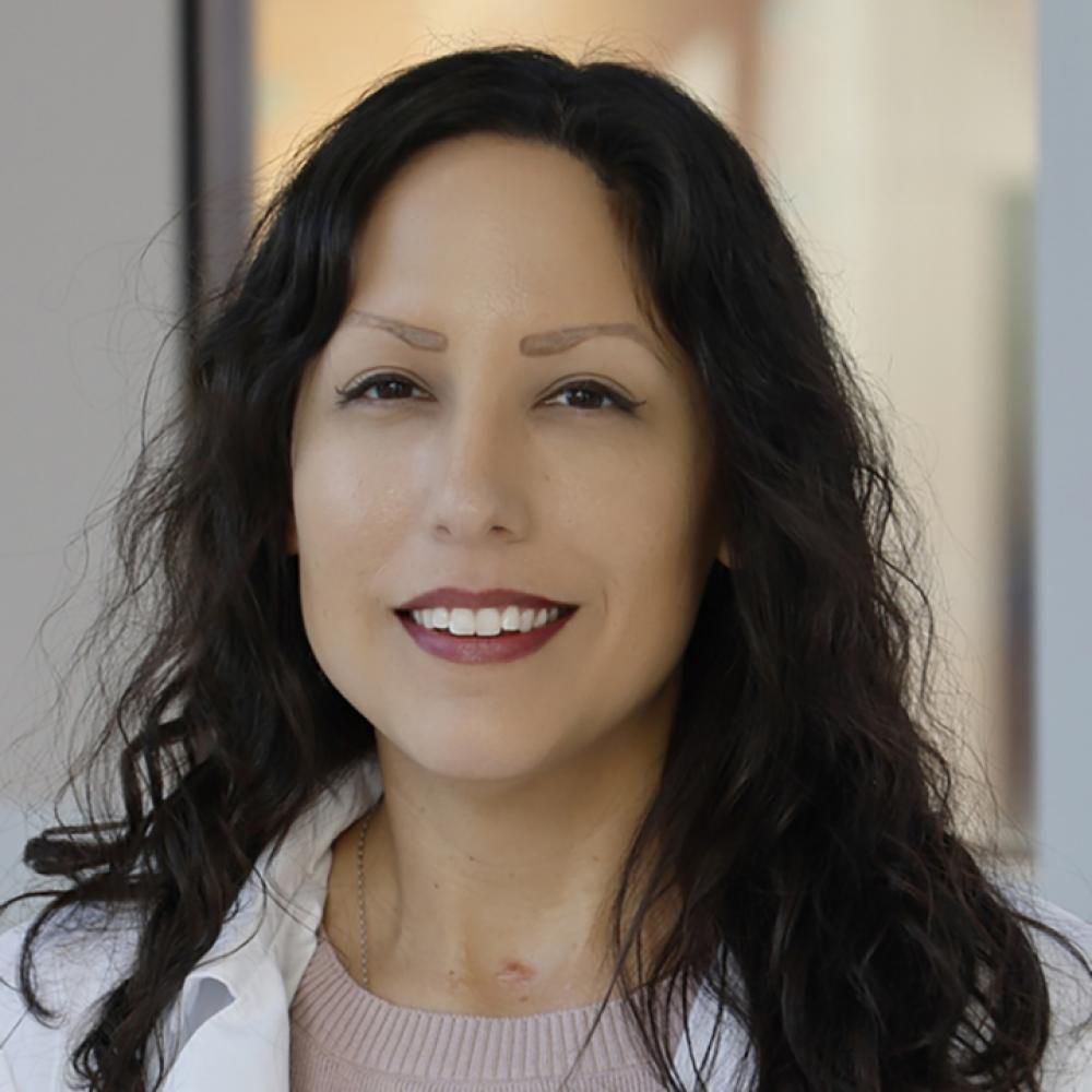 Headshot of Dr. Dionisia Quiroga. She is smiling facing forward in a clinical setting and has dark-brown hair at shoulder-length.