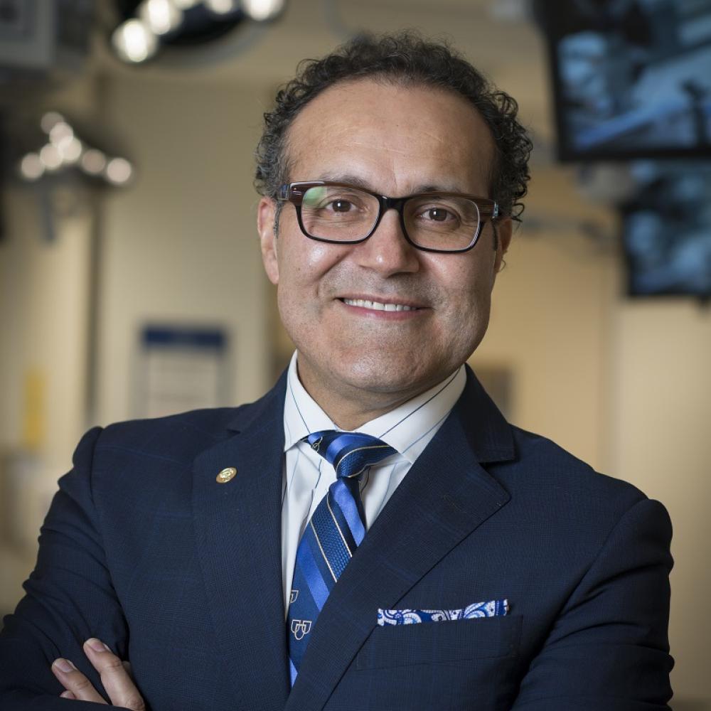 Headshot of Dr. Alfredo Quiñones-Hinojosa in a striped blue tie and navy blue jacket. He's wearing glasses and is smiling facing forward, and he's standing with arms crossed in a clinical setting.