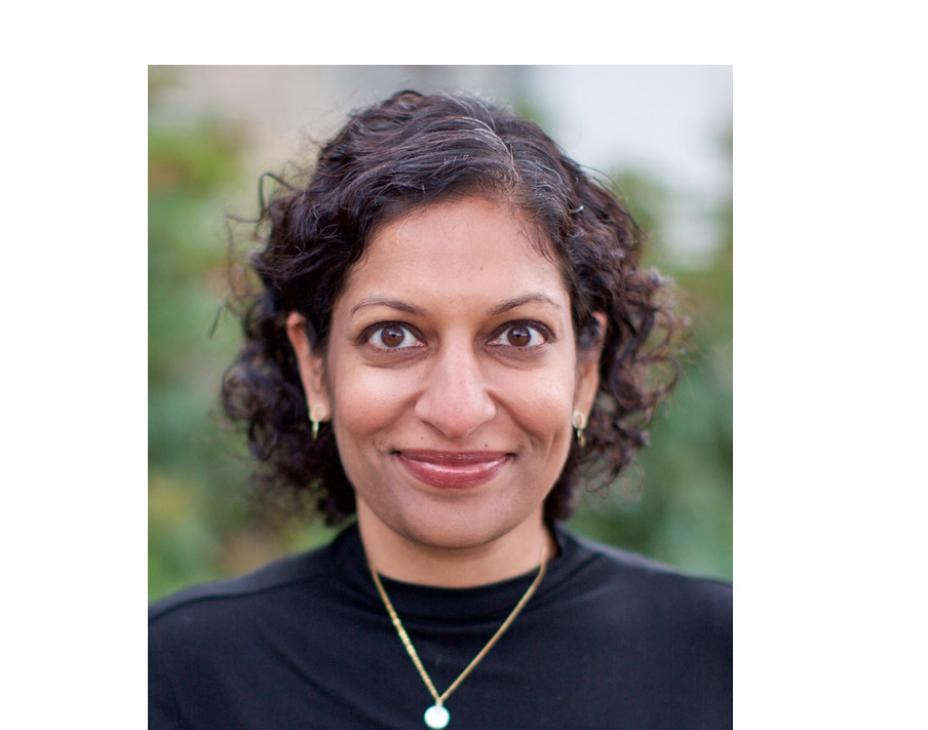 Dr. Veena Shankaran headshot. She is smiling facing forward in an outdoor setting. She has neck-length dark brown hair and is wearing a black sweater with a white necklace.