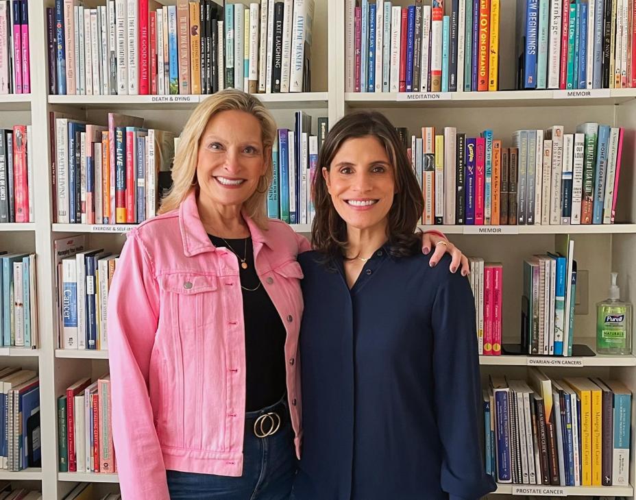 From left to right: Brenda and Stephanie. Brenda is wearing a pink jacket and blue jeans, and Stephanie is wearing a dark-blue shirt and blue jeans. Both are standing in front of a bookcase and smiling facing forward.