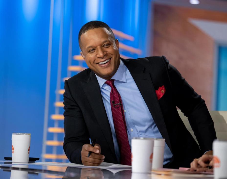 NBC journalist Craig Melvin sitting at his broadcast newsdesk, smiling facing to the side.