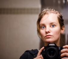 Anya Magnuson looking straight forward into a mirror while holding her camera.