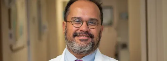 Dr. Flavio Rocha facing forward and smiling. He is wearing a white coat. He's in a well-lit hallway that appears to be in a hospital or clinic.