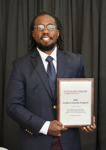 Dr. Terrell Coring holding his AMRA plaque at the 2022 ASCO Annual Meeting. He is smiling facing forward against a dark curtain, and is wearing a navy blue suit. His hair is black and shoulder-length.