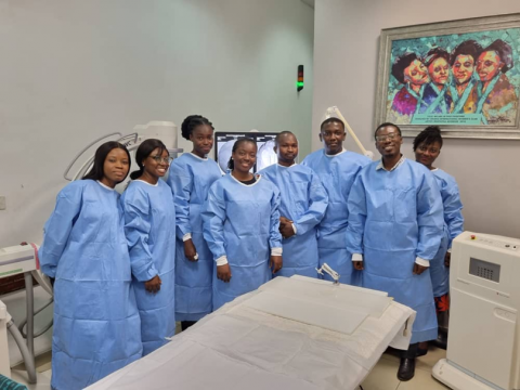 Dr. Aba Scott with seven members from her local cancer care team in Ghana. They are all wearing blue health-provider gowns and are standing together, smiling facing forward, in a clinic setting.