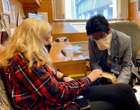 From left to right: Anya Magnuson and Dr. Jithma Abeykoon. Both are sitting in a clinical office and wearing masks. Dr. Abeykoon is observing Anya's hand, and Anya is looking down at her hand.