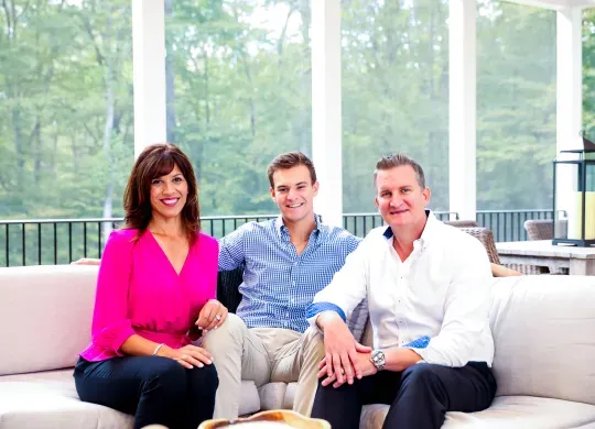 Left to right: Nancy Berry, Jake Berry, Alex Berry. All three are seated facing forward and smiling, located in a screened-in sun-room with white columns and green trees visible in the background. Nancy is wearing a bright pink shirt with dark blue pants. Jake is wearing a light blue shirt with khaki plants. Alex is wearing a white shirt with black pants.