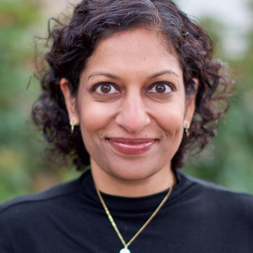 Dr. Veena Shankaran smiling facing forward in an outdoor setting. She is wearing a black shirt with a necklace, and has neck-length dark brown hair.