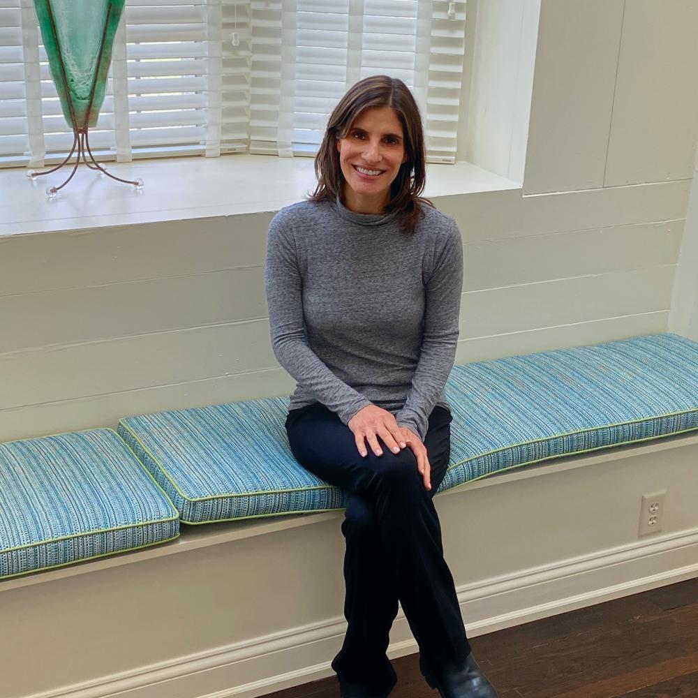 Stephanie Stern sitting cross-legged on a sofa at Hope Connections. She's wearing a gray turtleneck shirt and black pants/shoes, and smiling facing forward.