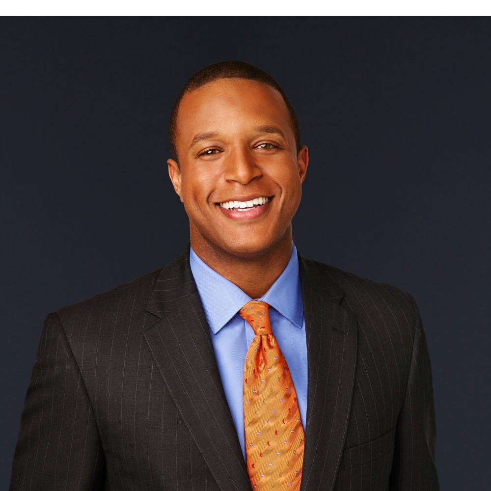 NBC journalist Craig Melvin smiling facing forward wearing a black suit with orange tie.