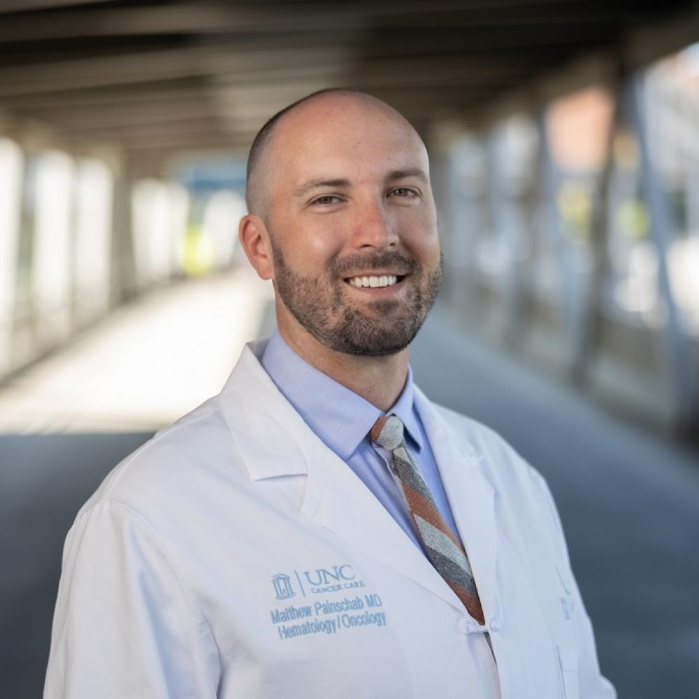 Dr. Matthew Painschab outdoors on a bridge wearing a white coat and smiling facing forward.