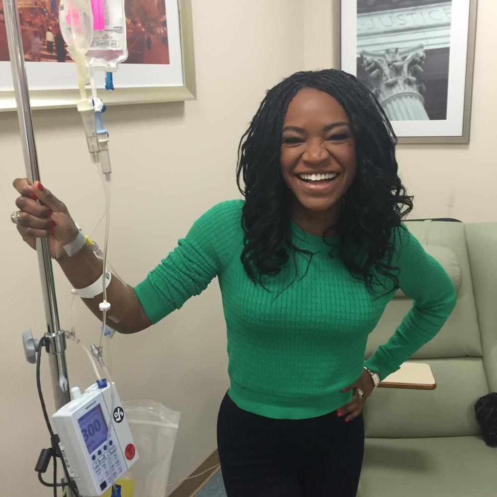 Athena Jones in a clinical setting. She is smiling, standing upright, facing forward with a hand on her hip, with an IV machine connected to her arm.